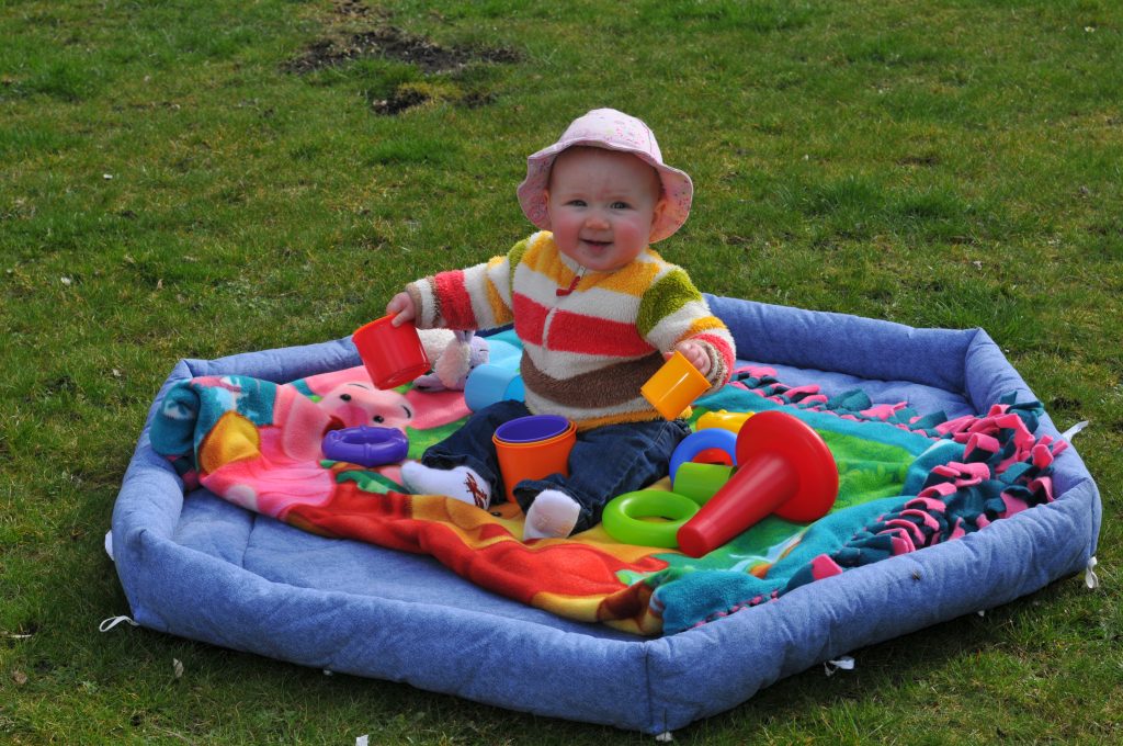 A baby sitting on a play mat, surounded by toys, on a grass lawn.